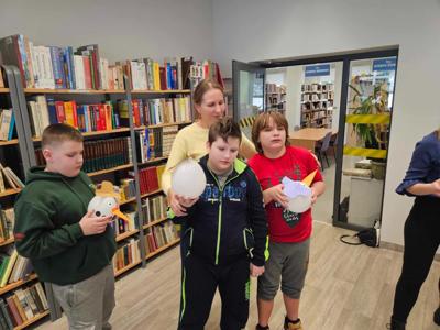 Na zdjęciu znajdują się uczestnicy zajęć. Dzieci stoją. Zdjęcie wykonano w bibliotece. W tle księgozbiór biblioteki.
