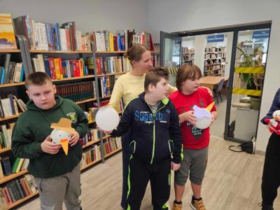Na zdjęciu znajdują się uczestnicy zajęć. Dzieci stoją. Zdjęcie wykonano w bibliotece. W tle księgozbiór biblioteki.