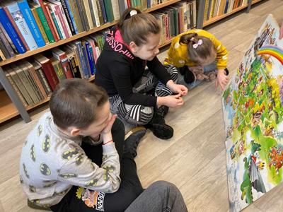 Na zdjęciu znajdują się uczestnicy zajęć. Dzieci siedzą na podłodze. Zdjęcie wykonano w bibliotece. W tle księgozbiór biblioteki.