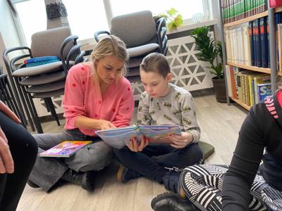 Na zdjęciu znajdują się uczestnicy zajęć. Dzieci siedzą na podłodze. Zdjęcie wykonano w bibliotece. W tle księgozbiór biblioteki.