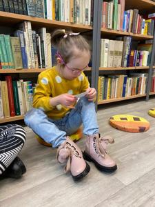 Na zdjęciu znajdują się uczestnicy zajęć. Dzieci siedzą na podłodze. Zdjęcie wykonano w bibliotece. W tle księgozbiór biblioteki.