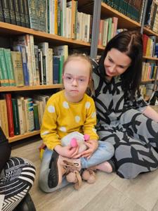 Na zdjęciu znajdują się uczestnicy zajęć. Dzieci siedzą na podłodze. Zdjęcie wykonano w bibliotece. W tle księgozbiór biblioteki.