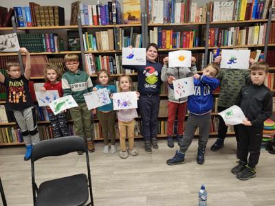 Na zdjęciu zdjęciu znajdują się uczestnicy warsztatów. Uczestnicy stoją i trzymają w ręce wykonane prace plastyczne. W tle księgozbiór biblioteki.
