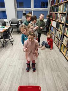 Na zdjęciu znajdują się uczestnicy warsztatów. Uczestnicy stoją. W tle księgozbiór biblioteki.