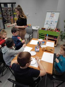 Na zdjęciu znajdują się uczestnicy warsztatów. Uczestnicy siedzą przy stołach. W tle księgozbiór biblioteki.