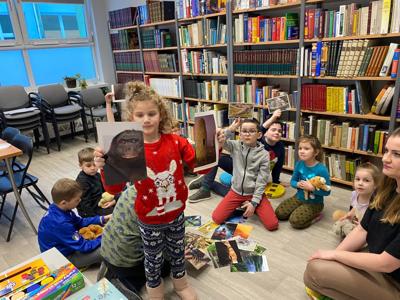 Na zdjęciu znajdują się uczestnicy warsztatów. Uczestnicy siedzą. W tle księgozbiór biblioteki.