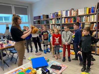 Na zdjęciu znajdują się uczestnicy warsztatów. Uczestnicy stoją. W tle księgozbiór biblioteki.
