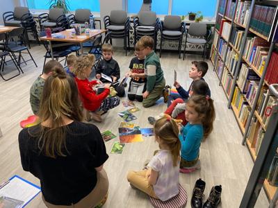 Na zdjęciu znajdują się uczestnicy warsztatów. Uczestnicy siedzą. W tle księgozbiór biblioteki.
