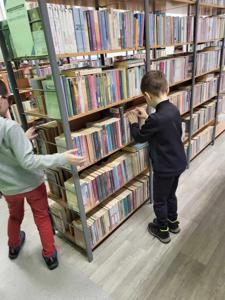 Na zdjęciu znajdują się uczestnicy warsztatów. Uczestnicy stoją. W tle księgozbiór biblioteki.