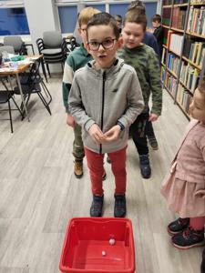 Na zdjęciu znajdują się uczestnicy warsztatów. Uczestnicy stoją. W tle księgozbiór biblioteki.