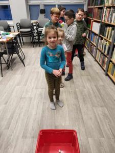 Na zdjęciu znajdują się uczestnicy warsztatów. Uczestnicy stoją. W tle księgozbiór biblioteki.