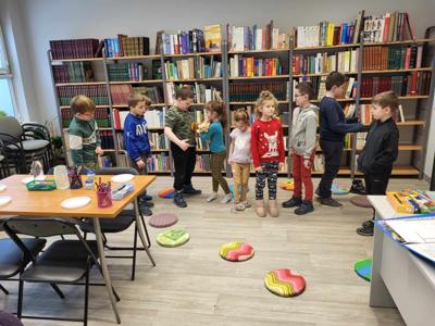 Na zdjęciu znajdują się uczestnicy warsztatów. Uczestnicy stoją. W tle księgozbiór biblioteki.