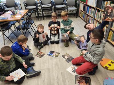Na zdjęciu znajdują się uczestnicy warsztatów. Uczestnicy siedzą. W tle księgozbiór biblioteki.