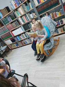 Na zdjęciu znajdują się uczestnicy zajęć. Siedzą na fotelu, dorosła osoba czyta książkę mając dziecko na kolanach. Zdjęcie wykonano w bibliotece.