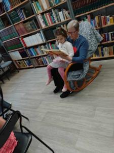Na zdjęciu znajdują się uczestnicy zajęć. Siedzą na fotelu, dorosła osoba czyta książkę mając dziecko na kolanach. Zdjęcie wykonano w bibliotece.