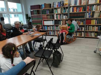Na zdjęciu znajdują się uczestnicy zajęć. Siedzą na fotelu, dorosła osoba czyta książkę mając dziecko na kolanach. Zdjęcie wykonano w bibliotece.