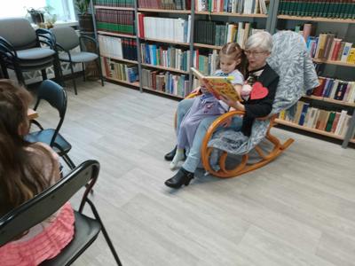Na zdjęciu znajdują się uczestnicy zajęć. Siedzą na fotelu, dorosła osoba czyta książkę mając dziecko na kolanach. Zdjęcie wykonano w bibliotece.