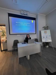 Na zdjęciu znajduje się prelegentka. W tle wyświetlana prezentacja multimedialna. Zdjęcie wykonano w bibliotece.