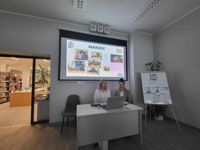 Na zdjęciu znajduje się prelegentki. W tle wyświetlana prezentacja multimedialna. Zdjęcie wykonano w bibliotece.