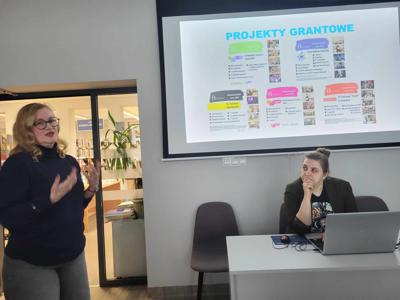 Na zdjęciu znajduje się prelegentki. W tle wyświetlana prezentacja multimedialna. Zdjęcie wykonano w bibliotece.
