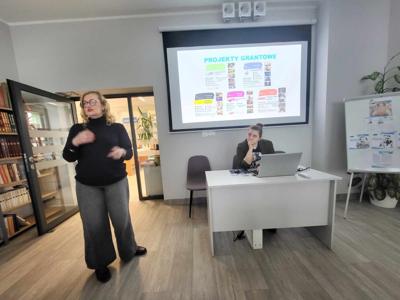 Na zdjęciu znajduje się prelegentki. W tle wyświetlana prezentacja multimedialna. Zdjęcie wykonano w bibliotece.