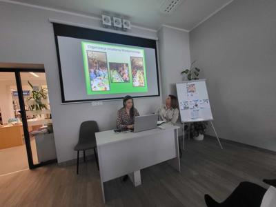 Na zdjęciu znajduje się prelegentki. W tle wyświetlana prezentacja multimedialna. Zdjęcie wykonano w bibliotece.