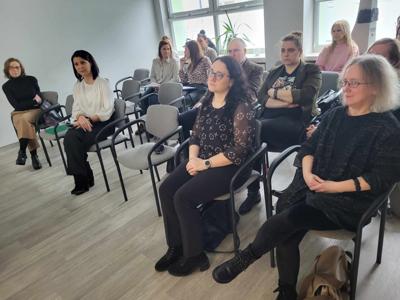 Na zdjęciu znajdują się osoby siedzące na krzesłach. W tle wyświetlana prezentacja multimedialna. Zdjęcie wykonano w bibliotece.