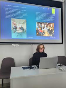 Na zdjęciu znajduje się prelegentka. W tle wyświetlana prezentacja multimedialna. Zdjęcie wykonano w bibliotece.