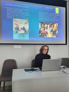 Na zdjęciu znajduje się prelegentka. W tle wyświetlana prezentacja multimedialna. Zdjęcie wykonano w bibliotece.