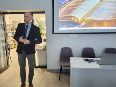 Na zdjęciu znajduje się prelegent. W tle wyświetlana prezentacja multimedialna. Zdjęcie wykonano w bibliotece.