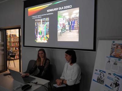 Na zdjęciu znajduje się prelegentki. W tle wyświetlana prezentacja multimedialna. Zdjęcie wykonano w bibliotece.