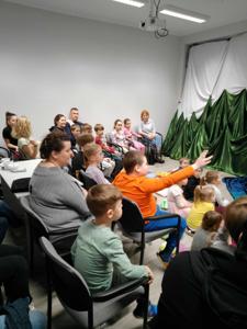 Uczestnicy siedzą na podłodze i na krzesłach. W tle teatrzyk i cienie.