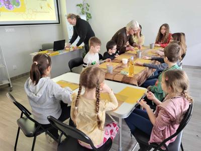 Na zdjęciu znajdują się uczestnicy warsztatów. Uczestnicy siedzą przy stołach i wykonują świece. Zdjęcie wykonano w bibliotece.