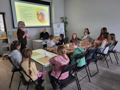 Na zdjęciu znajdują się uczestnicy warsztatów. Uczestnicy siedzą przy stołach i wykonują świece. Zdjęcie wykonano w bibliotece.