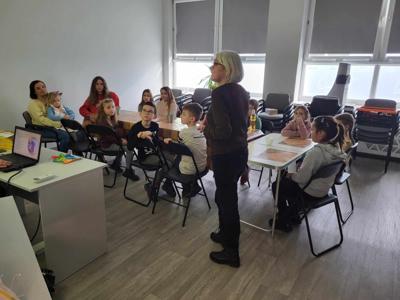 Na zdjęciu znajdują się uczestnicy warsztatów. Uczestnicy siedzą przy stołach i wykonują świece. Zdjęcie wykonano w bibliotece.