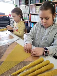 Na zdjęciu znajdują się uczestnicy warsztatów. Uczestnicy siedzą przy stołach i wykonują świece. Zdjęcie wykonano w bibliotece.