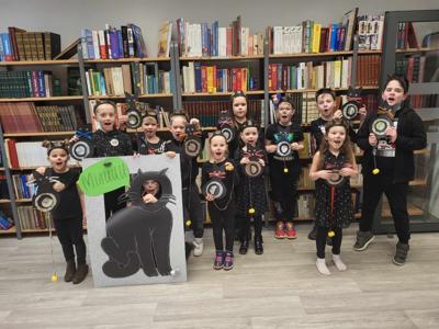 Na zdjęciu znajdują się uczestnicy warsztatów. Dzieci stoją i trzymają w rękach prace plastyczne - kotki. W tle księgozbiór biblioteki.