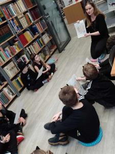 Na zdjęciu znajdują się uczestnicy zajęć siedzący na poduszkach. Zdjęcie wykonano w bibliotece.