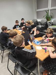 Na zdjęciu znajdują się uczestnicy zajęć siedzący przy stole. Dzieci wykonują prace plastyczne. Zdjęcie wykonano w bibliotece.