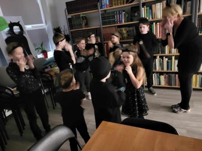 Na zdjęciu znajdują się uczestnicy zajęć. Dzieci stoją i pokazują. różne rzeczy. Zdjęcie wykonano w bibliotece.