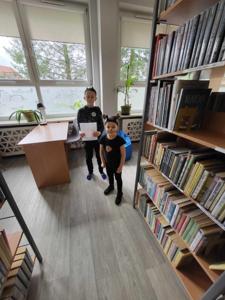 Na zdjęciu znajdują się uczestnicy zajęć. Dzieci stoją.  Zdjęcie wykonano w bibliotece.