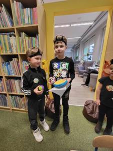 Na zdjęciu znajdują się uczestnicy zajęć. Dzieci stoją i trzymają kartki w rękach.  Zdjęcie wykonano w bibliotece.
