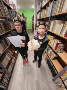 Na zdjęciu znajdują się uczestnicy zajęć. Dzieci stoją i trzymają kartki w rękach.  Zdjęcie wykonano w bibliotece.
