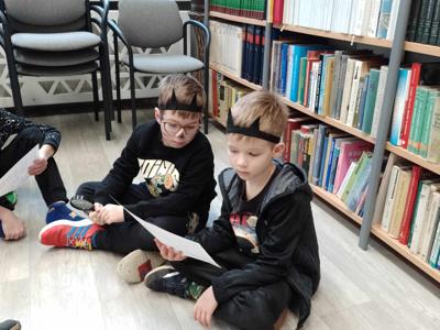 Na zdjęciu znajdują się uczestnicy zajęć siedzący na poduszkach. Zdjęcie wykonano w bibliotece.