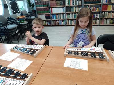 Na zdjęciu znajdują się uczestnicy zajęć. Dzieci siedzą przy stole i grają na cymbałkach.