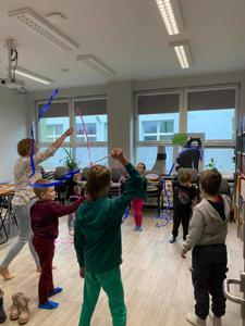 Na zdjęciu znajdują się uczestnicy zajęć. Dzieci stoją w kręgu i ruszają wstążkami.