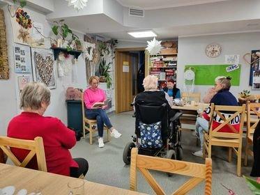 Zdjęcie przedstawia seniorów zgromadzonych przy stole. Seniorzy wsłuchani w czytaną książkę.  Zdjęcie wykonano w domu seniora.
