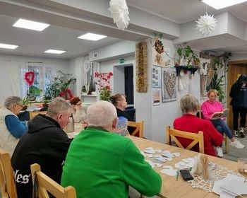 Zdjęcie przedstawia seniorów zgromadzonych przy stole. Seniorzy wsłuchani w czytaną książkę.  Zdjęcie wykonano w domu seniora.