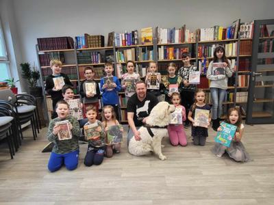 Na zdjęciu zgadują się uczestnicy zajęć. Na zdjęciu znajduje się pies. Uczestnicy trzymają w rękach książki. W tle księgozbiór biblioteki.