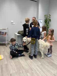.Na zdjęciu zgadują się uczestnicy zajęć. Na zdjęciu znajduje się pies. Uczestnicy siedzą oraz stoją głaskając psa.